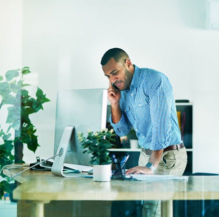 African-american-business-man-in-a-blue-shirt-standing-behind-his-computer-at-his-desk-while-on-the-phone.