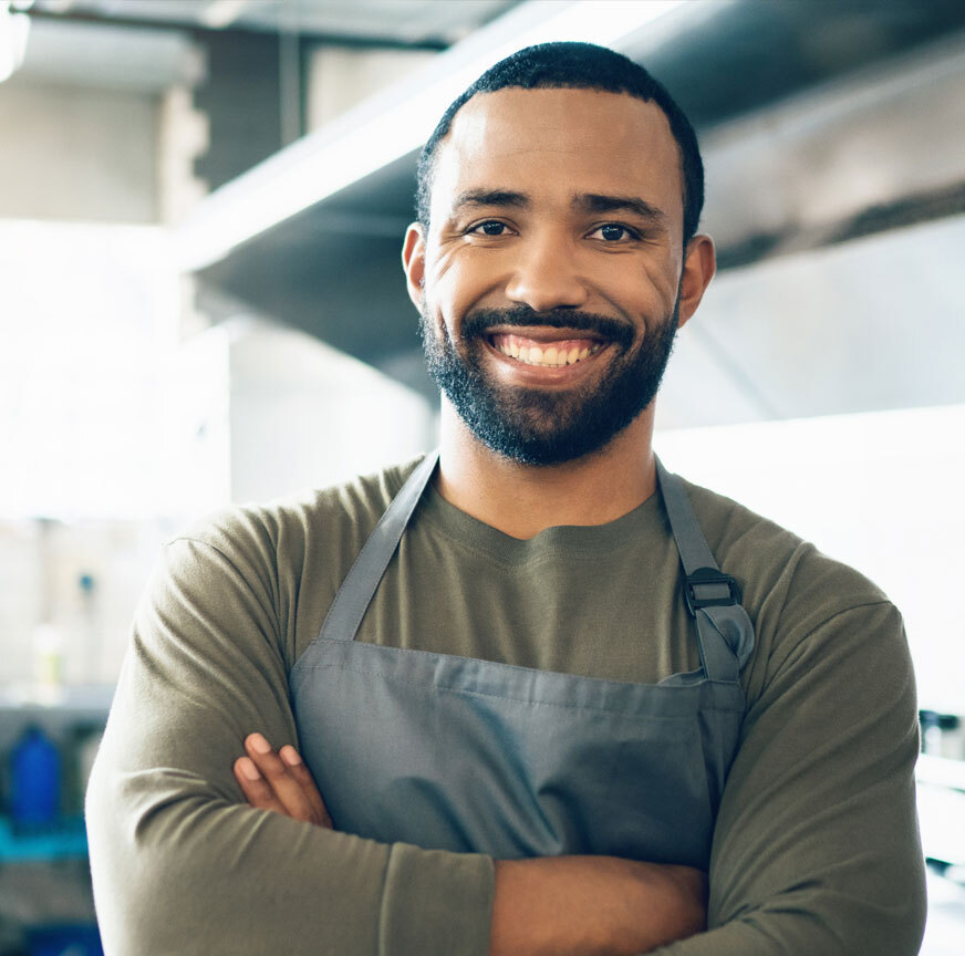 Business owner smiling standing with his arms crossed while wearing an apron in his restaurants kitchen.