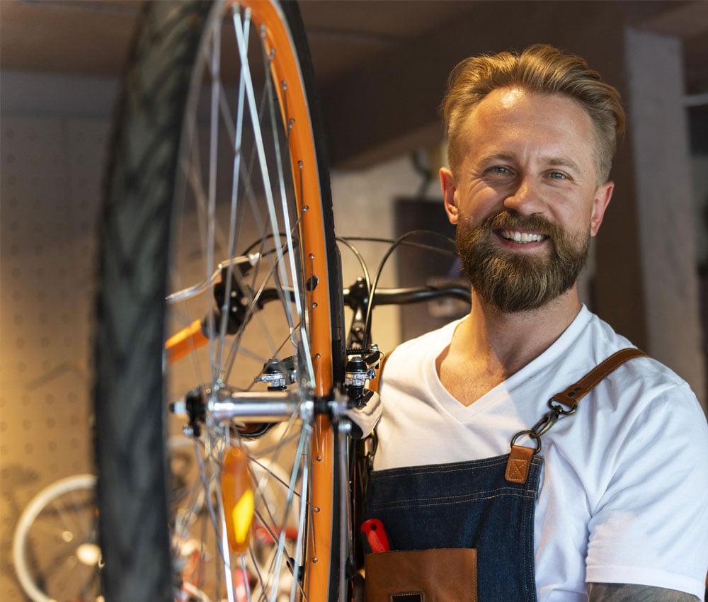 Smiling business owner of a bike shop wearing an apron and holding a bike wheel thinking about his Business Savings Account